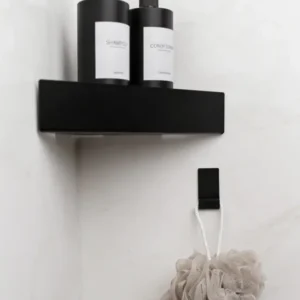 Minimalistisches schwarzes Eckregal aus schwarzem pulverbeschichteten Metall an einer weißen Wand mit zwei mattschwarzen Spenderflaschen für Shampoo und Conditioner. Darunter hängt ein beigefarbener Duschschwamm an einem passenden schwarzen Metall-Wandhaken TOULON ebenfalls pulverbeschichtet matt schwarz.