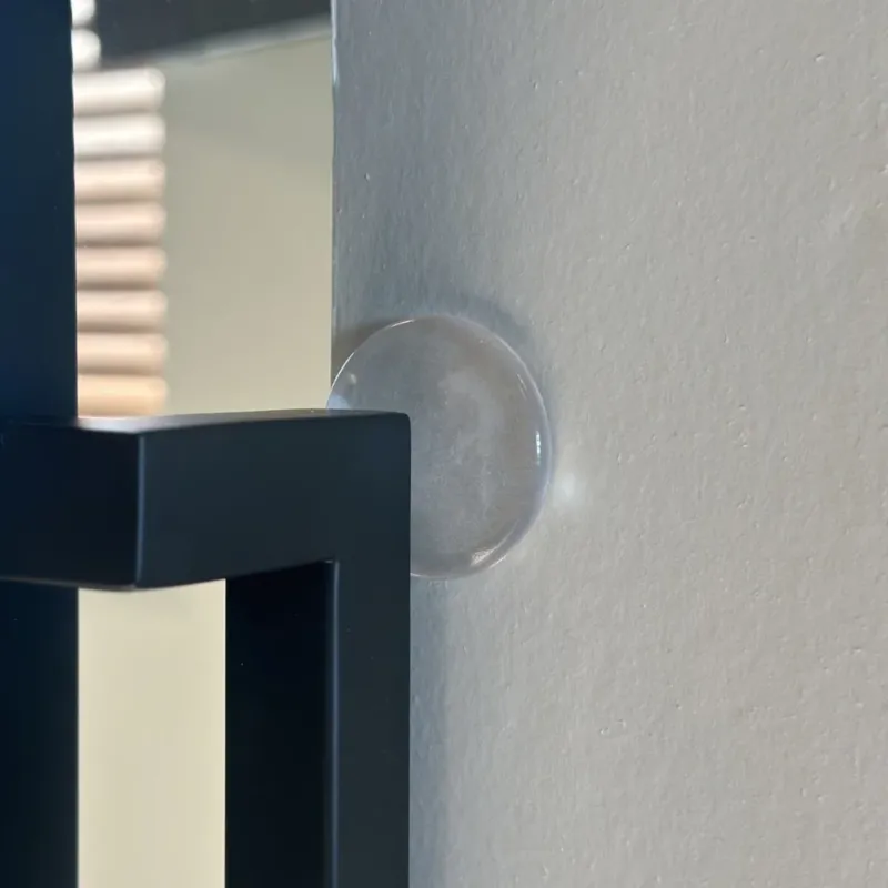 Der 6x selbstklebende Türstopper ⌀2cm von yessential.de haftet an einer beigen Wand, sodass ein schwarz umrandeter Spiegel die Oberfläche nicht berührt und beschädigt.
